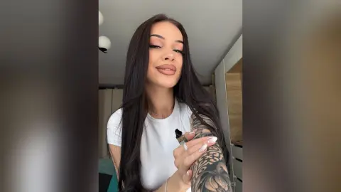 AriaVex poses with long dark hair and a white t-shirt, showing off her large arm tattoo.