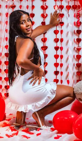 Woman in white summer dress posing against red heart decorations for a romantic Valentine's Day photoshoot.
