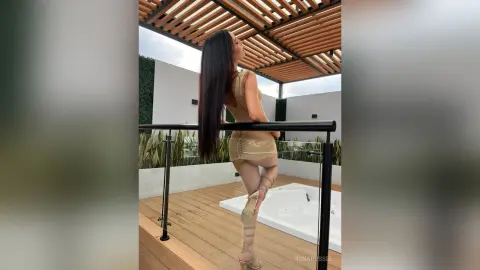 IrinaRussel poses sideways on a wooden deck, showing her long dark hair and gold outfit while standing near a hot tub under a slatted pergola.