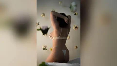 Woman in beige bikini posing amidst floating white flowers in a dreamy, ethereal indoor setting.