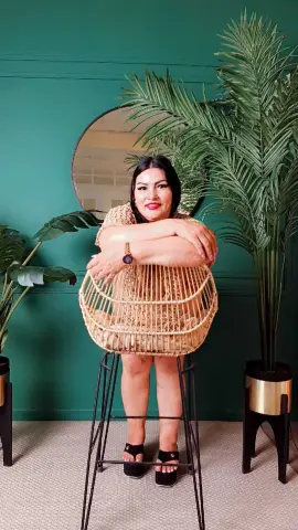 Woman in a beige knit top sitting on a rattan chair against a green wall with tropical palm plants.