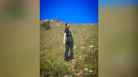 Young woman in black athletic wear standing in a sunny wildflower meadow under a clear blue sky.