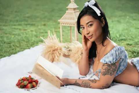 Young woman in a blue gingham sundress reading a book on a white picnic blanket in a sunny green meadow.