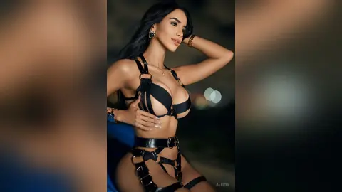 Alison poses in black harness-style lingerie with gold jewelry against a dark, blurred background.