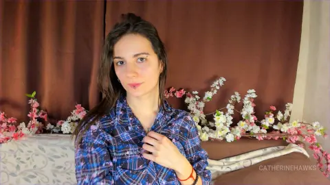 Young woman in a blue plaid shirt smiling in a cozy room with pink floral decorations.