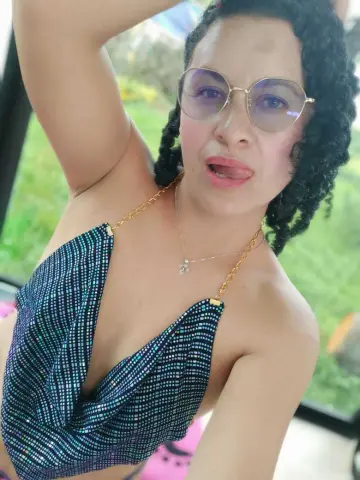Smiling woman in a sparkly blue halter top and glasses posing outdoors in a sunny garden setting.
