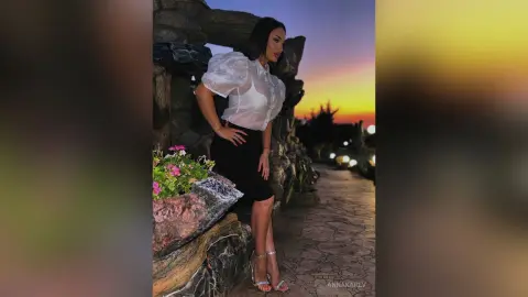 AnnaKarev poses in a white puffy blouse and black skirt outdoors at sunset.
