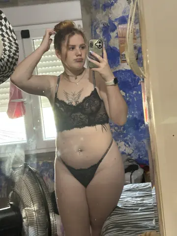 Young woman in black lace lingerie taking a mirror selfie in a sunlit bedroom.