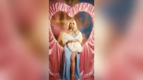 Glamorous blonde woman in a blue slit dress and white faux fur stole posing inside a pink heart frame.
