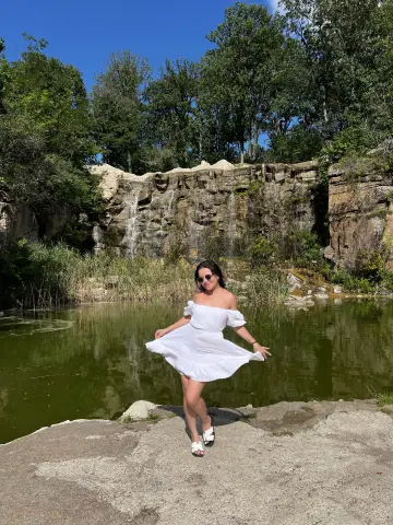 Woman in a white summer dress standing by a scenic waterfall and pond in a lush green forest.