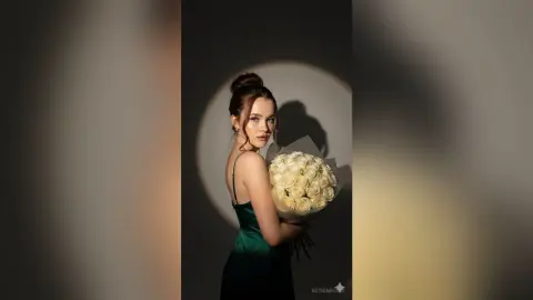 Elegant woman in a green satin dress holding a large bouquet of white roses in dramatic spotlight lighting.