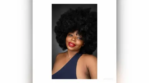 Smiling Black woman with voluminous afro hairstyle wearing a navy blue sleeveless top against a dark background.