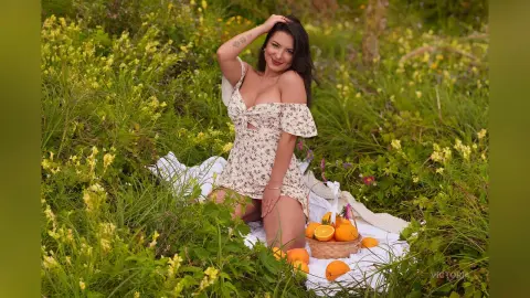 Smiling woman in a floral sundress sitting on a white blanket in a lush green meadow with oranges.