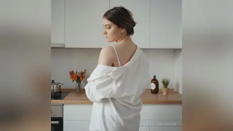 CarterMilena poses in a white shirt that hangs off her shoulders, showing her bare back and neck in a bright kitchen setting.