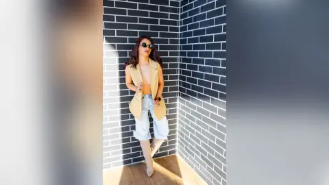 SofiaRoose poses in a tan vest and light blue denim shorts against a black brick wall, wearing sunglasses and letting her long dark hair hang loose.