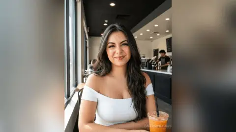 Smiling young woman in a white off-the-shoulder top sitting in a bright, modern cafe.