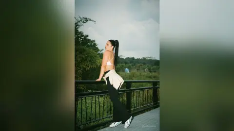 Young woman in white crop top and black leggings posing on a balcony overlooking a lush green forest.