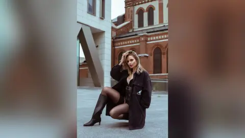 Gia poses in a black jacket and black lace lingerie while sitting on a concrete ledge outdoors.
