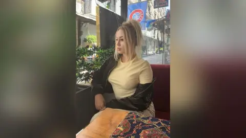 Blonde woman in a black jacket and cream top sitting by a window in a cozy cafe setting.