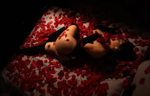 Woman in black bodysuit lying on a bed covered in red rose petals in a romantic, moody setting.