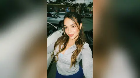 Smiling brunette woman in a grey zip-up hoodie posing outdoors in a parking lot at night.