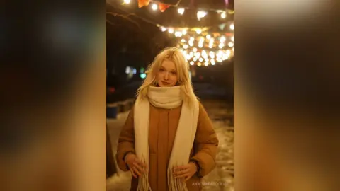 Blonde woman in a brown winter coat and white scarf smiling under warm festive string lights at night.