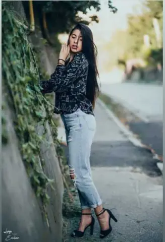 Young woman in a black patterned blouse and light blue jeans leaning against a green ivy wall outdoors.