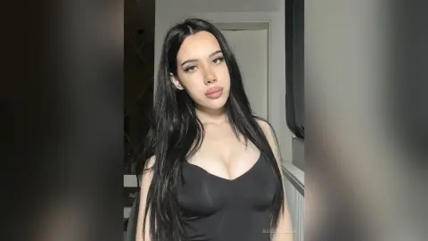 Young woman with long dark hair wearing a black tank top in a minimalist indoor setting with a moody vibe.