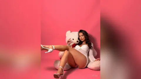 Glamorous woman in a white bodysuit and heels sitting by a giant teddy bear against a pink background.