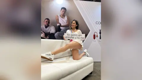 A woman in a white tank top and shorts sits on a white sofa holding a sign in a bright indoor setting.