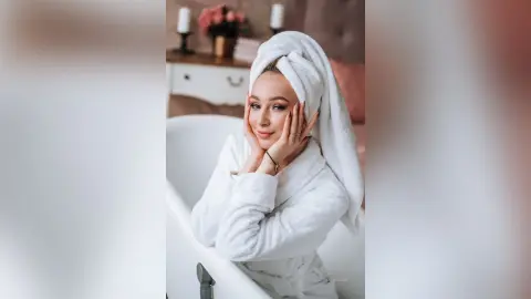 Smiling young woman in a white fluffy bathrobe and towel wrap relaxing in a bright, cozy bathroom setting.