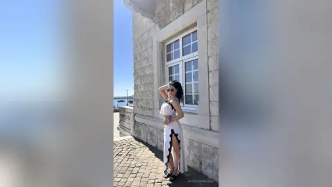 Stylish woman in a white maxi dress and sunglasses standing by a stone building on a sunny day.