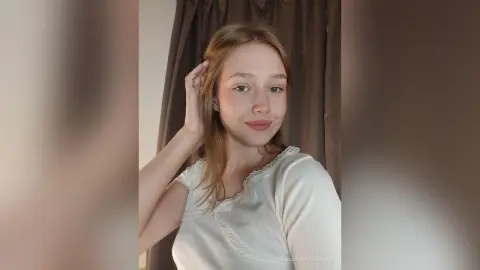 EvelinaMoorina is a young woman with long light brown hair wearing a white ribbed top, posing in front of brown curtains.
