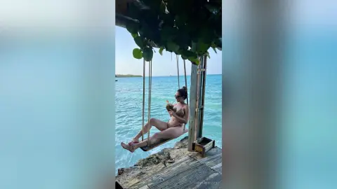 Woman in a bikini relaxing on a wooden swing over turquoise ocean water at a tropical beach resort.
