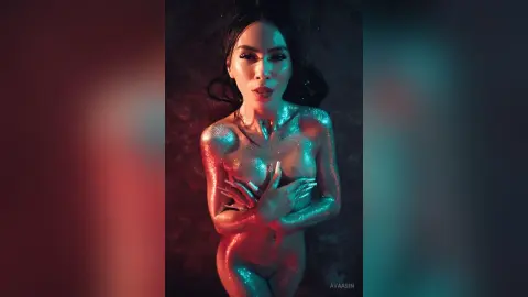 AvaaSin poses naked with her body covered in gold glitter, holding her hands over her breasts against a dark background.