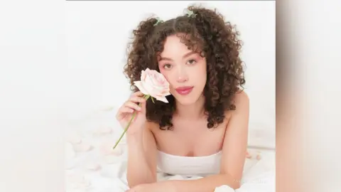 Young woman with curly hair holding a pink rose in a bright, dreamy aesthetic setting.