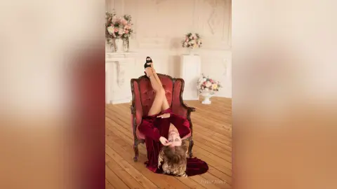 Elegant woman in a red velvet dress lounging playfully in a vintage armchair within a bright, floral studio.