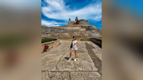 LauraRiveiro — LauraRiveiro stands on a stone walkway in a white crop top and shorts against a blue sky and old stone fortress.