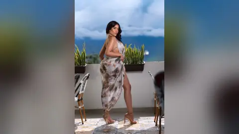 DayanaHille stands on a balcony wearing a long, patterned silk dress with a high slit, looking back over her shoulder against a blue sky.