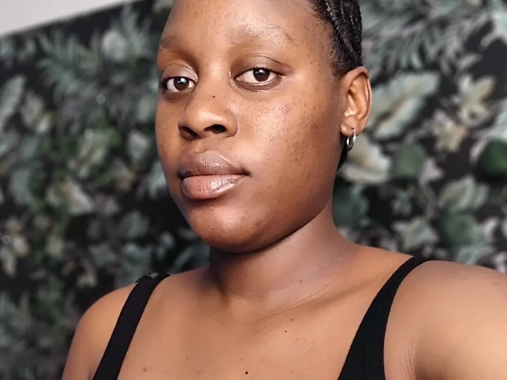 HaleyNoir looks directly at the camera wearing a black tank top, with her hair in tight braids against a dark leafy background.