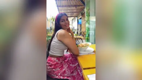 Smiling woman in a white tank top and pink floral skirt sitting at an outdoor cafe table.
