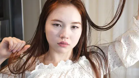 Young woman with long brown hair wearing a white eyelet blouse in a bright, airy indoor setting.