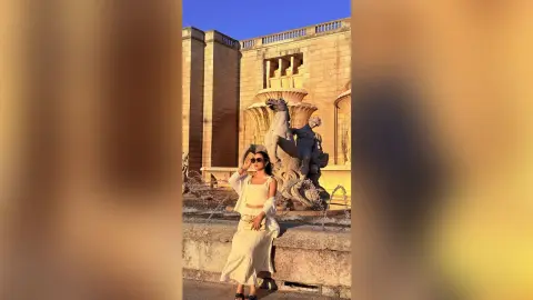 Stylish woman in white summer outfit posing by a stone fountain at a historic palace during golden hour.