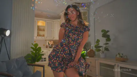 Smiling woman in a floral print dress playing with bubbles in a bright, cozy living room.