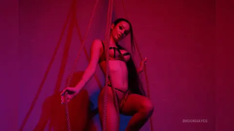 Young woman in black bikini posing between ropes under moody red lighting.