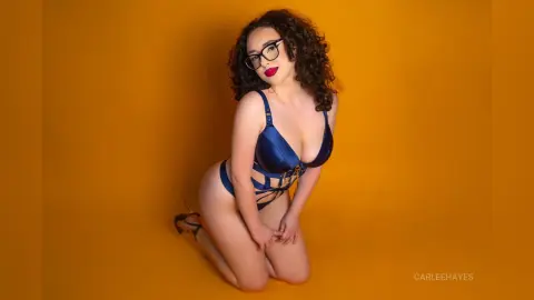Curvy woman with curly hair and glasses wearing navy blue lingerie against a vibrant yellow studio background.