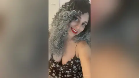Smiling woman with voluminous curly grey hair wearing a black floral print camisole in a bright indoor setting.