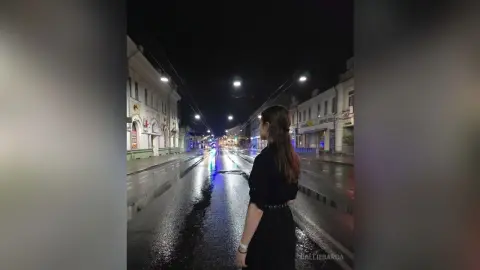 Young woman in a black dress walking down a wet city street at night under glowing streetlights.