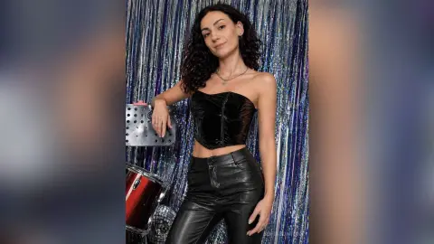 Stylish woman in a black velvet corset and leather pants posing against a shimmering silver tinsel backdrop.