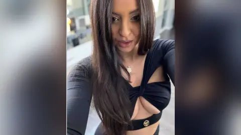 Brunette woman in a black long-sleeve crop top posing confidently in a bright indoor setting.